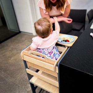May include: A light wood and black toddler learning tower with a child inside, reaching for food on a small tray. A woman with red hair is in the background. The tray has fruit and pancakes.