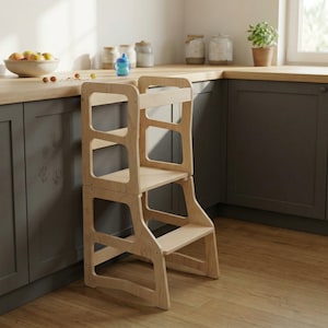 Puede incluir: Un taburete de cocina de madera clara con un diseño de dos escalones. El taburete tiene un marco rectangular con detalles recortados y un pequeño asa en la parte superior. Está diseñado para ayudar a los niños a alcanzar las encimeras.