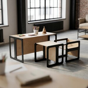 May include: A child's desk and chair set with a modern design. The desk and chair have a natural wood finish with black frames. The desk has a rectangular top and a lower shelf. The chair has a square seat and a backrest.