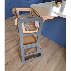 Puede incluir: Una torre de ayuda de cocina para niños pequeños de color gris y madera natural, con un arnés de seguridad y una pequeña mesa adjunta. La torre está diseñada para acercar a los niños a la altura de la encimera. La torre es de madera y tiene un marco gris.