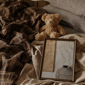 May include: A brown teddy bear sits on a bed with brown plaid bedding and a dark brown gingham pillow. A rectangular picture frame rests on the bed, displaying an image of a dark ceramic vase with thin branches against a textured wall.