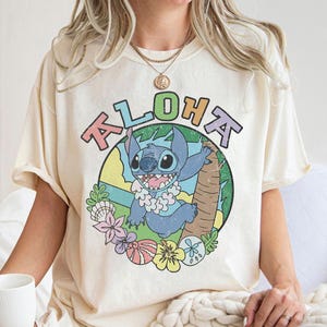 May include: Cream-colored t-shirt featuring a colorful graphic of Stitch from Lilo & Stitch, with the word "Aloha" above. The design includes a cartoon Stitch character, flowers, and palm trees. The shirt has short sleeves.