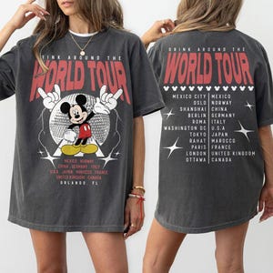 May include: Dark gray t-shirt with a graphic of Mickey Mouse and the text "DRINK AROUND THE WORLD TOUR" in red. The shirt lists cities like Mexico City, Berlin, and Tokyo. The design is on the front and back.