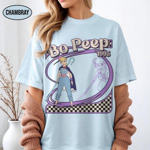 Peut inclure: T-shirt bleu clair avec un motif de Bo Peep de Toy Story, avec le texte "Bo Peep: 1995". Le motif comprend une illustration de dessin anim&eacute; de Bo Peep avec son b&acirc;ton et sa cape, sur un fond &agrave; carreaux violets et noirs.