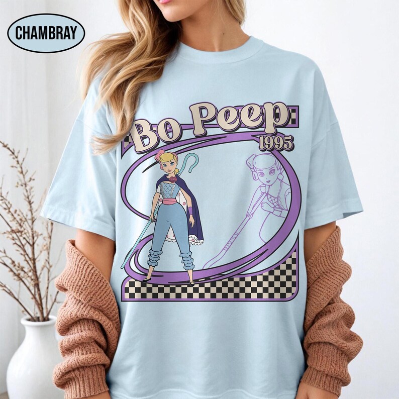 Peut inclure: T-shirt bleu clair avec un motif de Bo Peep de Toy Story, avec le texte "Bo Peep: 1995". Le motif comprend une illustration de dessin anim&eacute; de Bo Peep avec son b&acirc;ton et sa cape, sur un fond &agrave; carreaux violets et noirs.