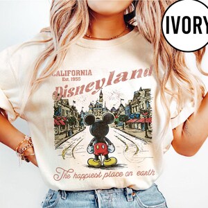 May include: Ivory-colored t-shirt featuring a vintage Disneyland graphic. The design includes Mickey Mouse, a street scene, and the text "California Disneyland Est. 1955" and "The happiest place on earth."