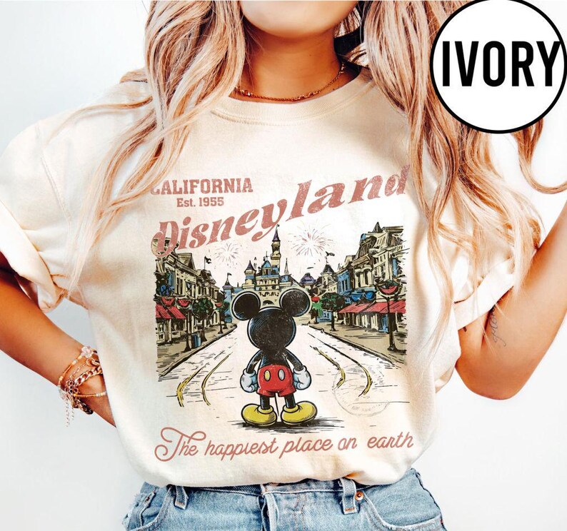 May include: Ivory-colored t-shirt featuring a vintage Disneyland graphic. The design includes Mickey Mouse, a street scene, and the text "California Disneyland Est. 1955" and "The happiest place on earth."