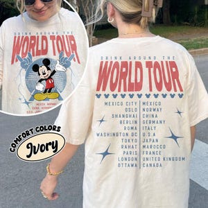 May include: Ivory-colored t-shirt with a graphic featuring Mickey Mouse and the text "DRINK AROUND THE WORLD TOUR." The shirt lists various cities. The front of the shirt has a circular graphic with Mickey Mouse. The back of the shirt also has the text "DRINK AROUND THE WORLD TOUR."