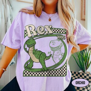 Peut inclure: T-shirt lilas avec un motif de dinosaure vert et le mot "Rex" dans une typographie r&eacute;tro. Le design comprend l'ann&eacute;e "1995" et un motif &agrave; carreaux. Le t-shirt a une coupe ample.