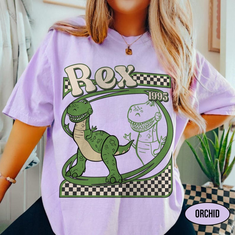 Peut inclure: T-shirt lilas avec un motif de dinosaure vert et le mot "Rex" dans une typographie r&eacute;tro. Le design comprend l'ann&eacute;e "1995" et un motif &agrave; carreaux. Le t-shirt a une coupe ample.