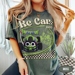 Peut inclure: T-shirt vert mousse avec un dessin de voiture de course verte et le texte "Re Car 1995". La voiture de course a de grands yeux de dessin anim&eacute; et un motif de drapeau &agrave; damier en bas. Le t-shirt est de coupe d&eacute;contract&eacute;e.