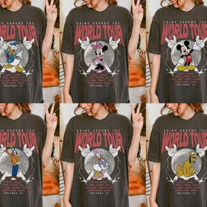 May include: Dark gray t-shirts with various Disney character graphics. Each shirt features the text "DRINK AROUND THE WORLD TOUR" in red, with a character like Donald Duck, Mickey Mouse, or Goofy in a disco ball design. The shirts also list cities like Mexico, Italy, and Orlando, FL.