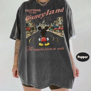 May include: Dark gray t-shirt featuring a graphic of Mickey Mouse standing on a road in front of Disneyland. The shirt has the text "CALIFORNIA Disneyland Est. 1955" and "The happiest place on earth" in a vintage style.