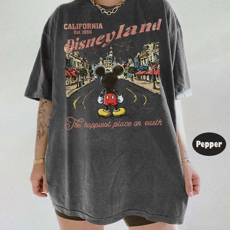 May include: Dark gray t-shirt featuring a graphic of Mickey Mouse standing on a road in front of Disneyland. The shirt has the text "CALIFORNIA Disneyland Est. 1955" and "The happiest place on earth" in a vintage style.