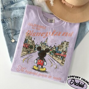 May include: A light purple t-shirt with a graphic of Mickey Mouse in front of Disneyland. The shirt has the text "CALIFORNIA Disneyland Est. 1955" and "The happiest place on earth". The shirt is made by Comfort Colors.