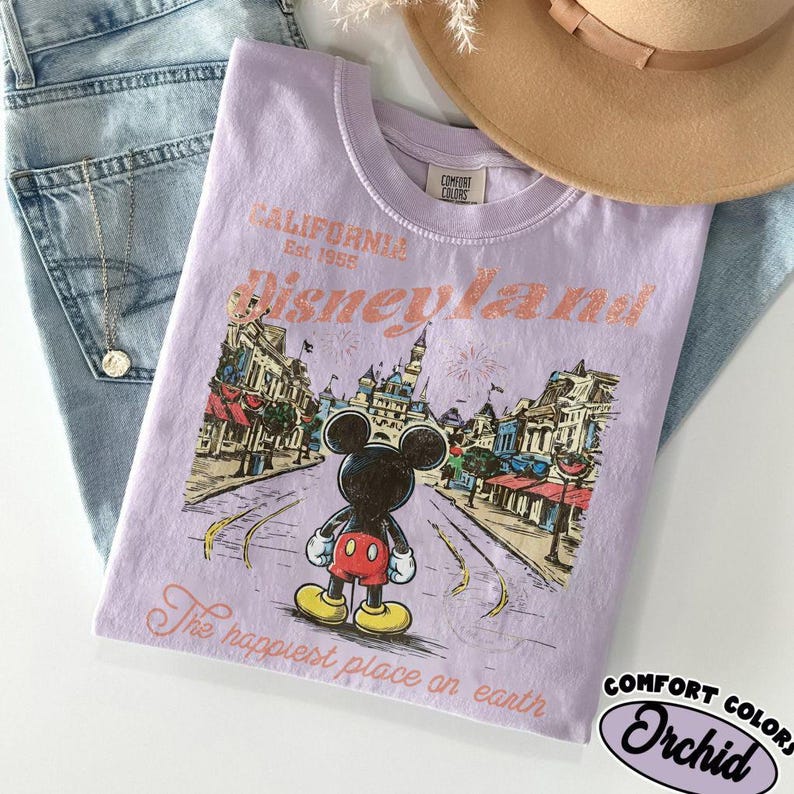 May include: A light purple t-shirt with a graphic of Mickey Mouse in front of Disneyland. The shirt has the text "CALIFORNIA Disneyland Est. 1955" and "The happiest place on earth". The shirt is made by Comfort Colors.