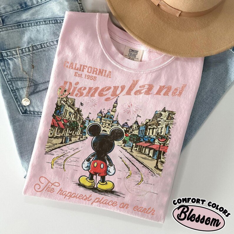 May include: A light pink t-shirt with a Disneyland graphic featuring Mickey Mouse. The shirt includes the text "California Est. 1955" and "The happiest place on earth". A tan hat and blue jeans are also in the image.