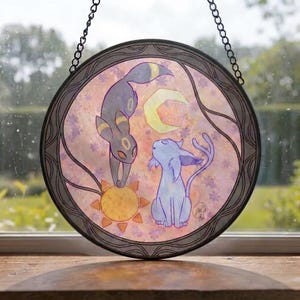 May include: A circular hanging suncatcher featuring a dark gray creature with yellow markings reaching for a yellow sun, and a light blue creature looking up at a yellow crescent moon, all set against a soft pink and purple floral background.