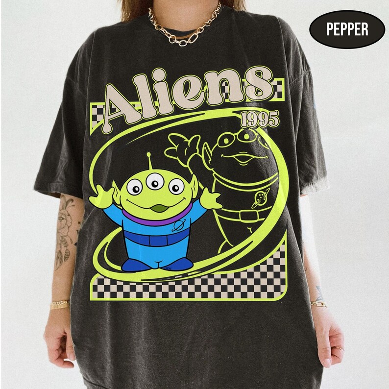 Peut inclure: T-shirt gris fonc&eacute; avec un dessin anim&eacute; de deux extraterrestres et le texte "Aliens 1995". Le motif comprend un extraterrestre vert &agrave; trois yeux et une tenue bleue et violette, et un second extraterrestre dans un ovale &agrave; carreaux noir et blanc.