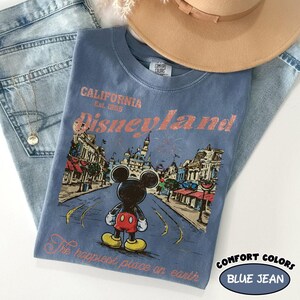 May include: Blue Comfort Colors t-shirt with a Disneyland graphic. The graphic features Mickey Mouse, a street scene, and the text "California Disneyland Est. 1955" and "The happiest place on earth". The shirt is paired with blue jeans and a tan hat.