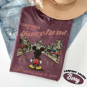 May include: Berry-colored t-shirt with a Disneyland graphic featuring Mickey Mouse. The shirt has the text "California Est. 1955 Disneyland" and "The happiest place on earth". A tan hat and blue jeans are in the background.