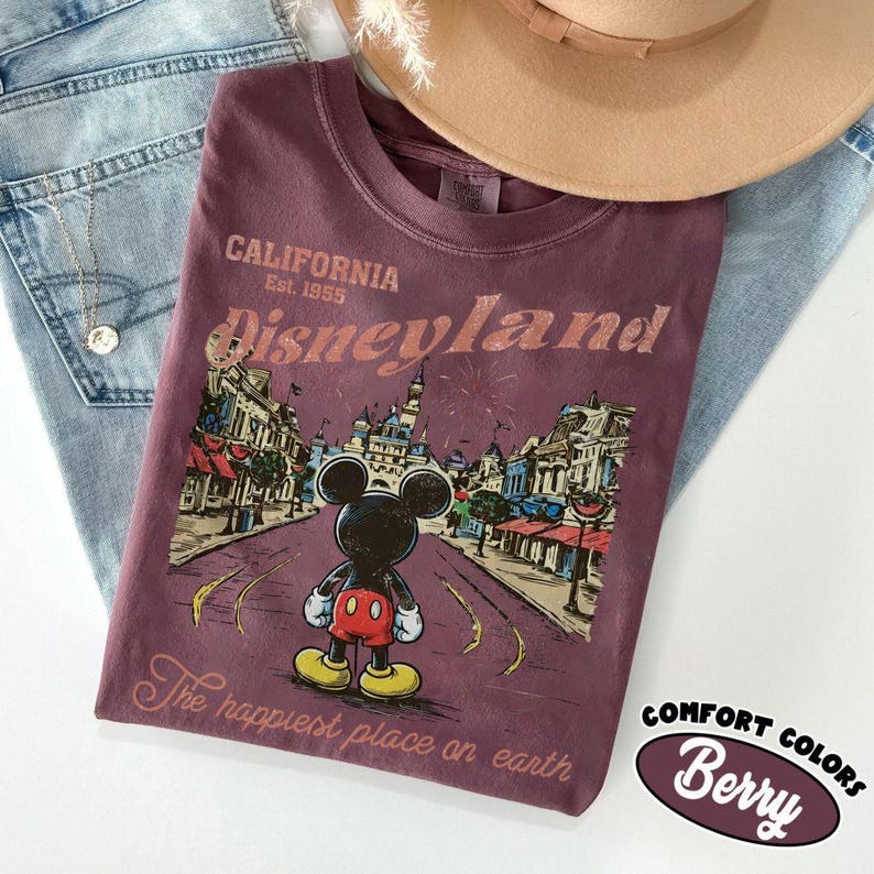 May include: Berry-colored t-shirt with a Disneyland graphic featuring Mickey Mouse. The shirt has the text "California Est. 1955 Disneyland" and "The happiest place on earth". A tan hat and blue jeans are in the background.