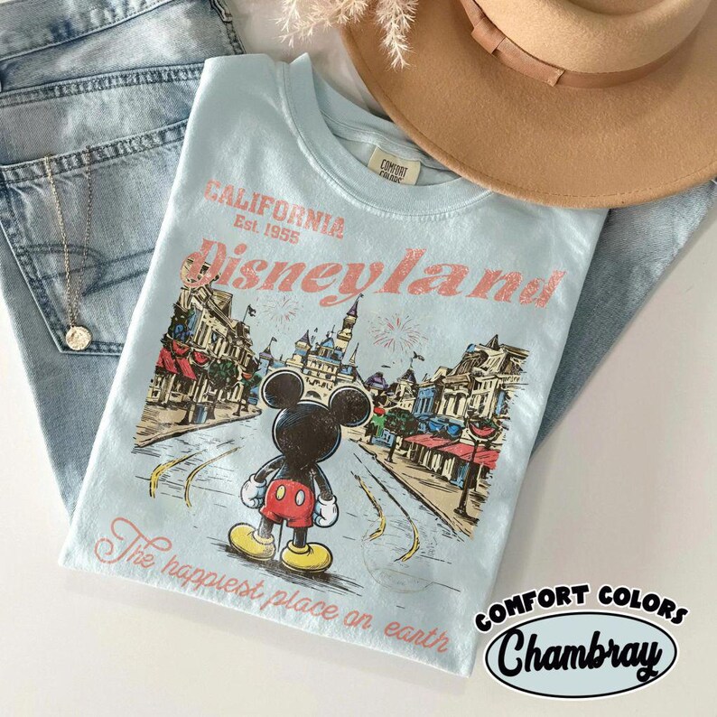 May include: Light blue t-shirt with a Disneyland graphic featuring Mickey Mouse. The shirt has the text "California Est. 1955 Disneyland" and "The happiest place on earth". The shirt is paired with blue jeans and a tan hat.