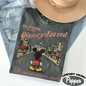 May include: A gray t-shirt with a Disneyland graphic featuring Mickey Mouse, a castle, and the text "California Est. 1955 Disneyland". The shirt is paired with blue jeans and a tan hat. The text "The happiest place on earth" is also printed on the shirt.