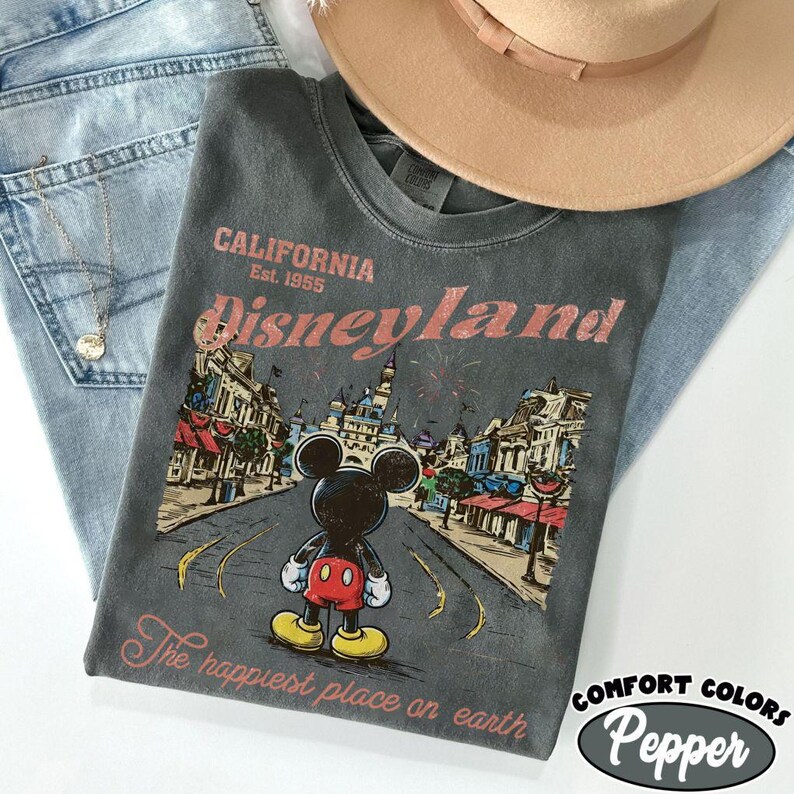 May include: A gray t-shirt with a Disneyland graphic featuring Mickey Mouse, a castle, and the text "California Est. 1955 Disneyland". The shirt is paired with blue jeans and a tan hat. The text "The happiest place on earth" is also printed on the shirt.