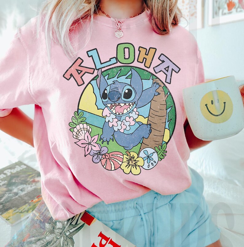 May include: Pink t-shirt with the word "ALOHA" above a cartoon character, Stitch, in a tropical scene. The character is wearing a flower lei. A white mug with a yellow smiley face is held in the hand. The shirt is paired with light blue shorts.