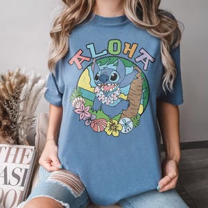 May include: Blue t-shirt featuring a cartoon image of Stitch from Lilo & Stitch. The design includes the word "ALOHA" in colorful letters, a smiling Stitch wearing a lei, and tropical flowers. The shirt is a comfortable, casual style.