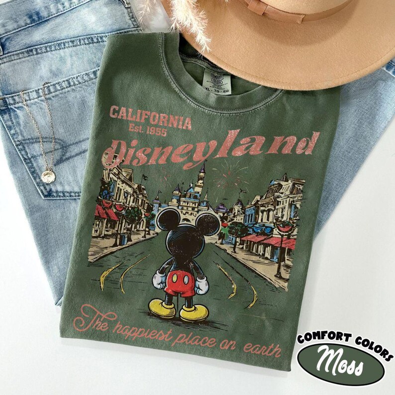 May include: Green t-shirt with a Disneyland graphic featuring Mickey Mouse, a castle, and the text "California Est. 1955". The shirt is paired with light blue jeans and a tan hat. The words "The happiest place on earth" are also printed on the shirt.