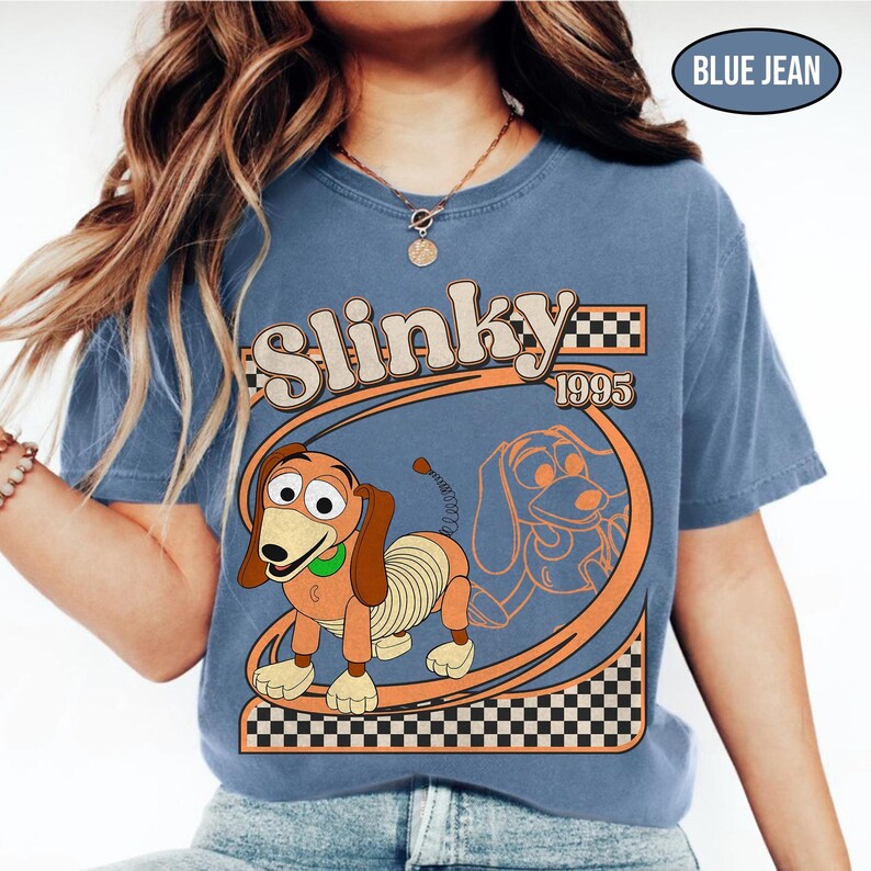 Peut inclure: T-shirt bleu jean avec une image de dessin anim&eacute; de Slinky Dog et le texte "Slinky 1995". Le design comprend un ovale orange et un motif &agrave; carreaux.