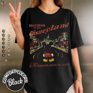 May include: Black t-shirt with a vintage Disneyland graphic. The design features Mickey Mouse standing on a road with buildings and the text "CALIFORNIA Disneyland Est. 1955" and "The happiest place on earth".