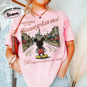 May include: Pink t-shirt with a Disneyland graphic featuring Mickey Mouse in front of a park scene. The shirt includes the text "California Est. 1955" and "The happiest place on earth". The shirt is labeled "Comfort Colors Blossom".