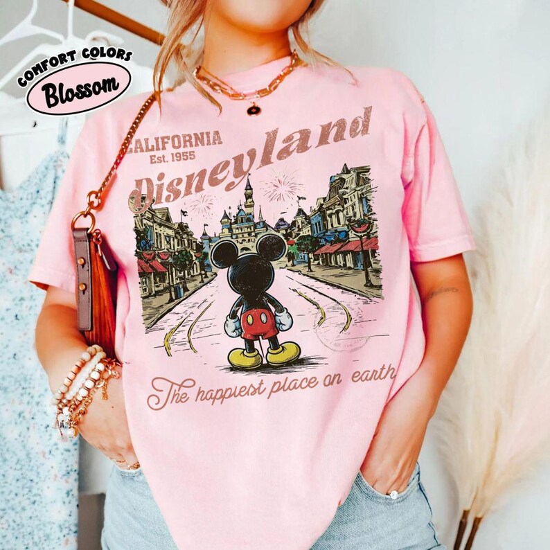May include: Pink t-shirt with a Disneyland graphic featuring Mickey Mouse in front of a park scene. The shirt includes the text "California Est. 1955" and "The happiest place on earth". The shirt is labeled "Comfort Colors Blossom".