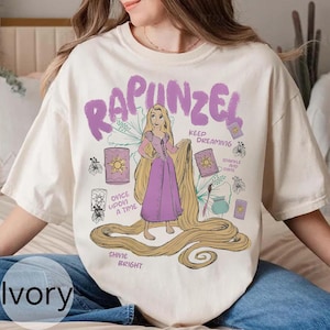 May include: Ivory-colored t-shirt featuring a graphic of Rapunzel with long blonde hair and a purple dress. The design includes the word "Rapunzel" in purple, along with the phrases "Once Upon a Time", "Keep Dreaming", and "Sparkle and Shine".