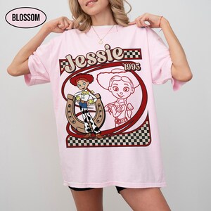 Peut inclure: T-shirt rose clair avec un motif de Jessie, personnage de Toy Story. Le design comprend le nom "Jessie", l'ann&eacute;e "1995" et une image de Jessie avec un fer &agrave; cheval. Le t-shirt est ample.