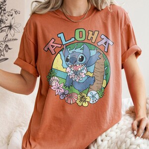 May include: Burnt orange t-shirt featuring a cartoon character with the word "ALOHA" above it. The character is blue with a flower lei and is set against a tropical background with flowers and a palm tree.