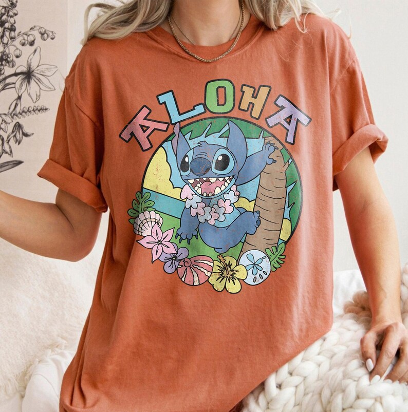 May include: Burnt orange t-shirt featuring a cartoon character with the word "ALOHA" above it. The character is blue with a flower lei and is set against a tropical background with flowers and a palm tree.