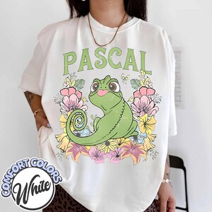 May include: White t-shirt featuring a green cartoon chameleon named Pascal, encircled by flowers. The word "PASCAL" is printed above the chameleon in green. The shirt has the words "COMFORT COLORS" and "White" printed on the bottom left.