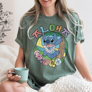 May include: Green t-shirt with the word "ALOHA" above a cartoon image of Stitch from Lilo & Stitch. The image features Stitch wearing a lei and surrounded by tropical flowers and plants. The shirt has rolled-up sleeves.