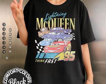Puede incluir: Camiseta negra con un gráfico de Rayo McQueen y otros personajes de Cars. El diseño incluye el texto "Lightning McQueen", "95" y "Think Fast". La camiseta es de Comfort Colors.