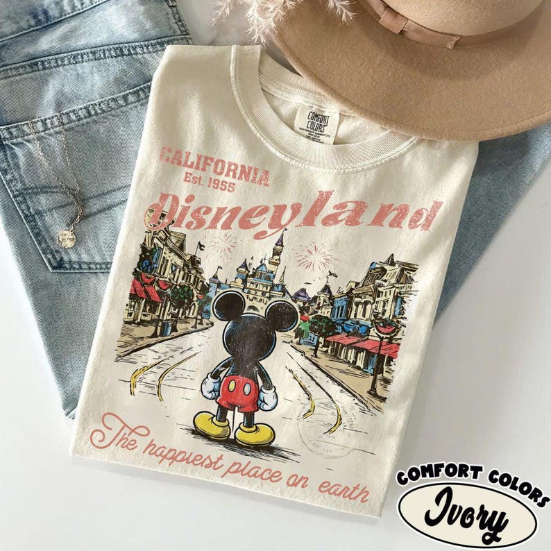 May include: Ivory-colored t-shirt with a Disneyland graphic featuring Mickey Mouse. The design includes the text "CALIFORNIA Est. 1955 Disneyland" and "The happiest place on earth". The shirt is from Comfort Colors.