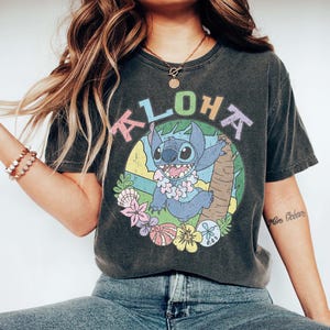 May include: Dark gray t-shirt featuring a colorful graphic of Stitch from Lilo & Stitch, with the word "ALOHA" above him. The design includes a tropical scene with flowers and a palm tree. The shirt is a relaxed fit.