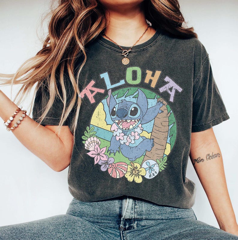 May include: Dark gray t-shirt featuring a colorful graphic of Stitch from Lilo & Stitch, with the word "ALOHA" above him. The design includes a tropical scene with flowers and a palm tree. The shirt is a relaxed fit.