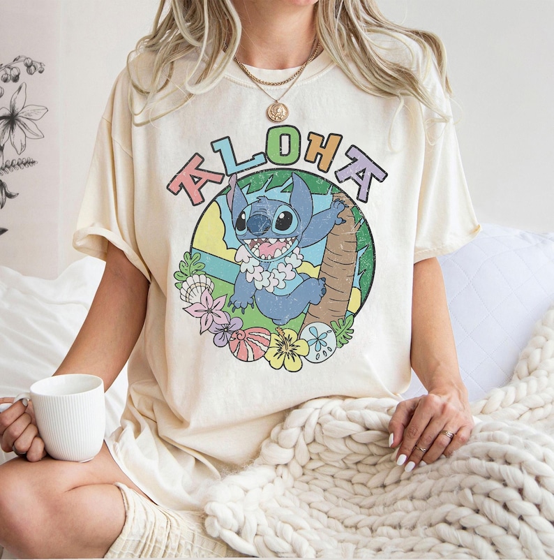 May include: Cream-colored t-shirt featuring a colorful graphic of Stitch from Lilo & Stitch, with the word "Aloha" above. The design includes a cartoon Stitch character, flowers, and palm trees. The shirt has short sleeves.