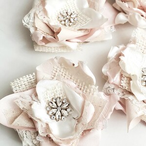 May include: A collection of handcrafted fabric flowers in shades of pale pink and cream. Each flower features layered satin and burlap petals, delicate white lace trim, and a central cluster of clear, faceted rhinestones arranged in a star shape.