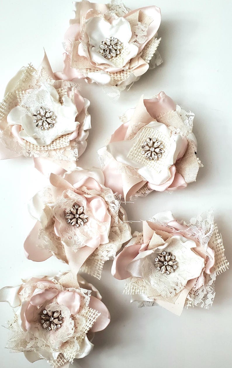 small fabric flowers made with bits of burlap, cotton, and lace. the center is accented by rhinestone small buttons. blush and ivory colors