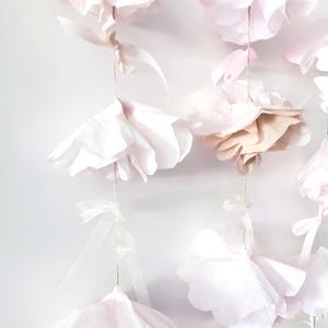 May include: A delicate garland of paper flowers in varying shades of pink and cream. The flowers are suspended by thin strings and adorned with small ribbons. The soft, layered petals create a romantic and airy aesthetic, perfect for event decor.